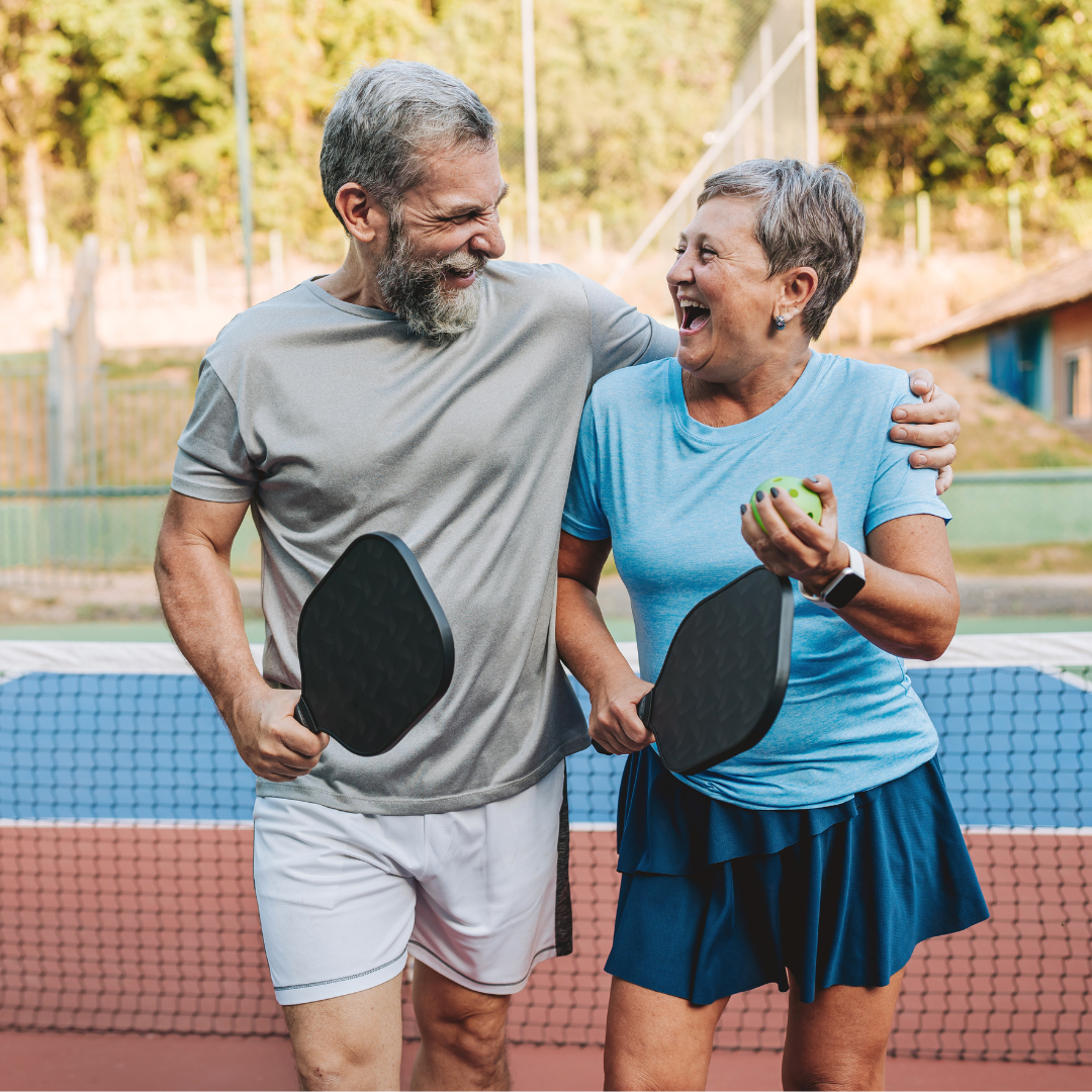 Two senior citizens look at one another smiling holding pickleball rackets.