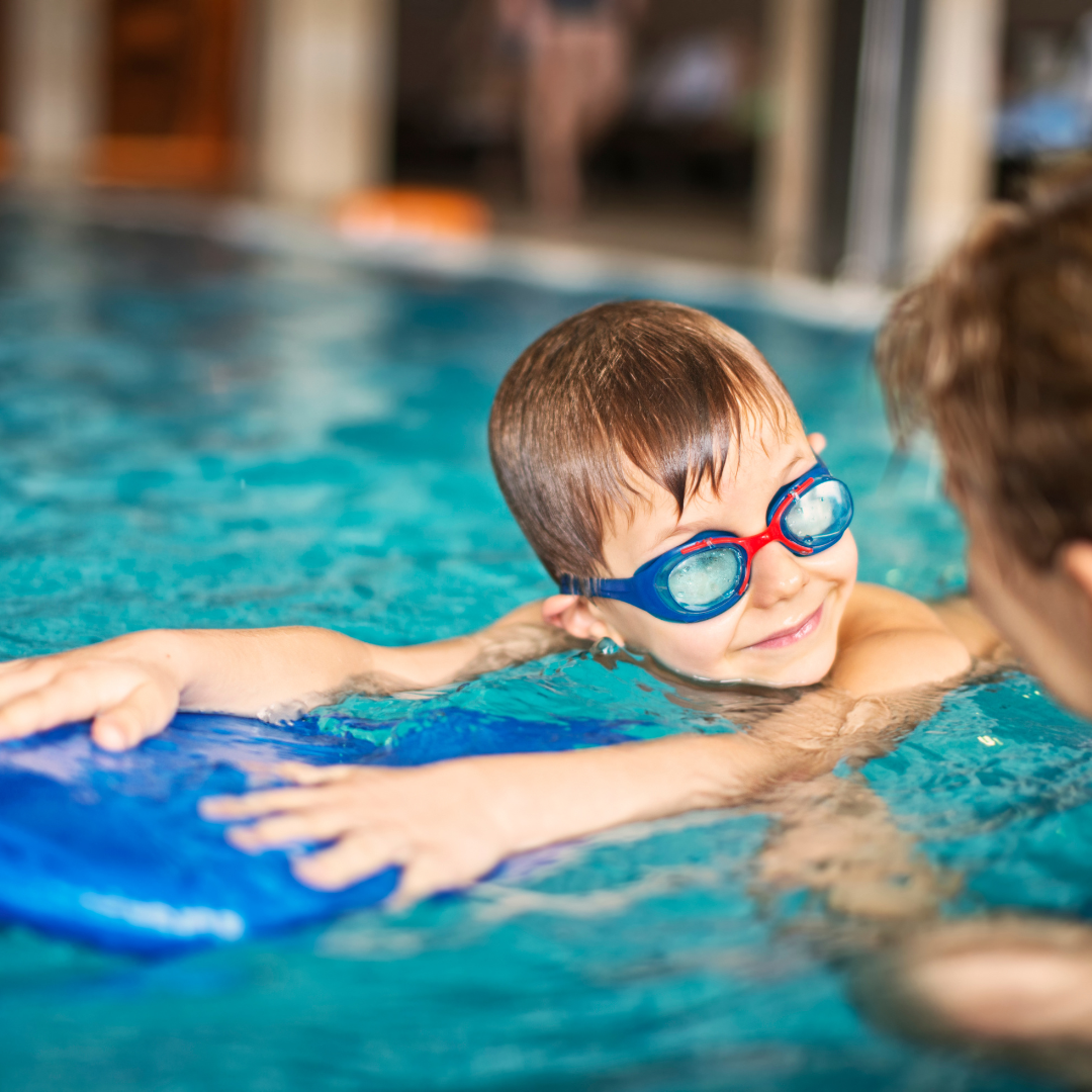 Child wearing goggles in a pool with a floatation device and adult nearby.